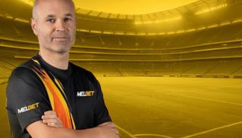 Andrés Iniesta wearing a MelBet jersey stands in a golden-lit football stadium, representing his new role as MelBet brand ambassador.