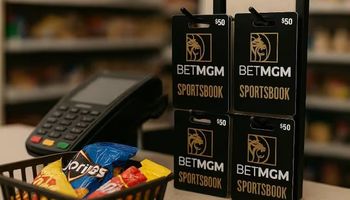 A realistic photo of a retail checkout counter with a POS terminal, a full shopping basket of snacks, and four BetMGM Sportsbook gift cards hanging on hooks behind the register.