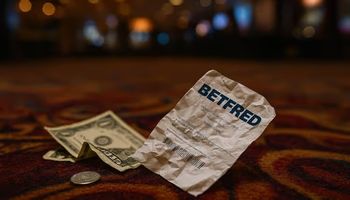 A crumpled Betfred sportsbook betting slip lies naturally on a casino floor, surrounded by a few scattered U.S. dollar bills and a quarter.
