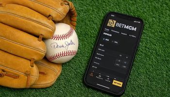 A realistic photo of a worn tan leather baseball glove holding a signed Derek Jeter baseball, placed beside a smartphone displaying the BetMGM sportsbook app on green turf.