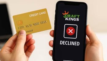 A person holding a credit card in front of a smartphone showing the DraftKings app with a red declined payment symbol.
