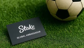 A business card with the Stake logo and “Global Ambassador” title lying on green football turf beside a vintage soccer ball.