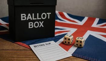 A black ballot box, ivory dice, and a folded betting slip on a wooden table partially covered by a Union Jack flag