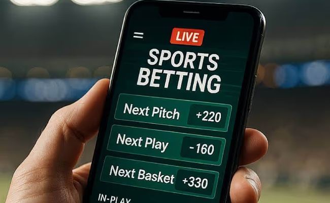 A smartphone held in a person’s hand displaying live microbetting options such as next pitch and next play, with a blurred stadium in the background.