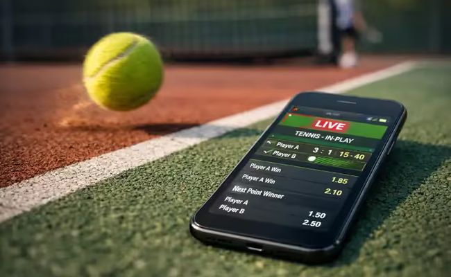 Smartphone displaying live tennis in-play betting interface on a tennis court with a ball landing out of bounds, symbolising illegal online wagering.
