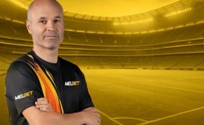 Andrés Iniesta wearing a MelBet jersey stands in a golden-lit football stadium, representing his new role as MelBet brand ambassador.